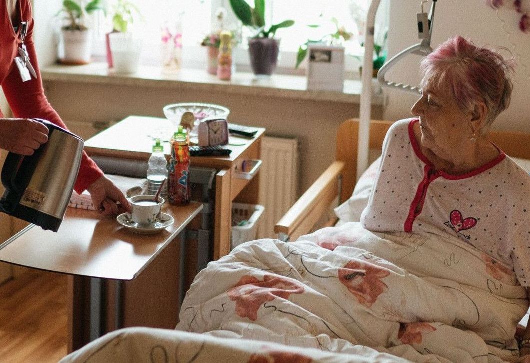 A caregiver serves tea to a senior woman in a cozy bedroom setting, fostering comfort and care.